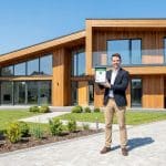 Un homme en tenue de ville tient une tablette devant une maison moderne en bois avec une déclaration d'achèvement validée.