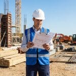 Chef de chantier en casque et gilet réfléchissant consultant des documents administratifs sur un site en construction.