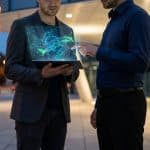 Deux hommes en tenue professionnelle examinant une interface holographique numérique au-dessus d'une tablette.