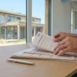 Mains remplissant un formulaire Cerfa de permis de construire avec une maison moderne en bois en arrière-plan.